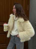 Woman holding a coffee cup, wearing a cream faux fur jacket over a white turtleneck sweater and grey jeans.