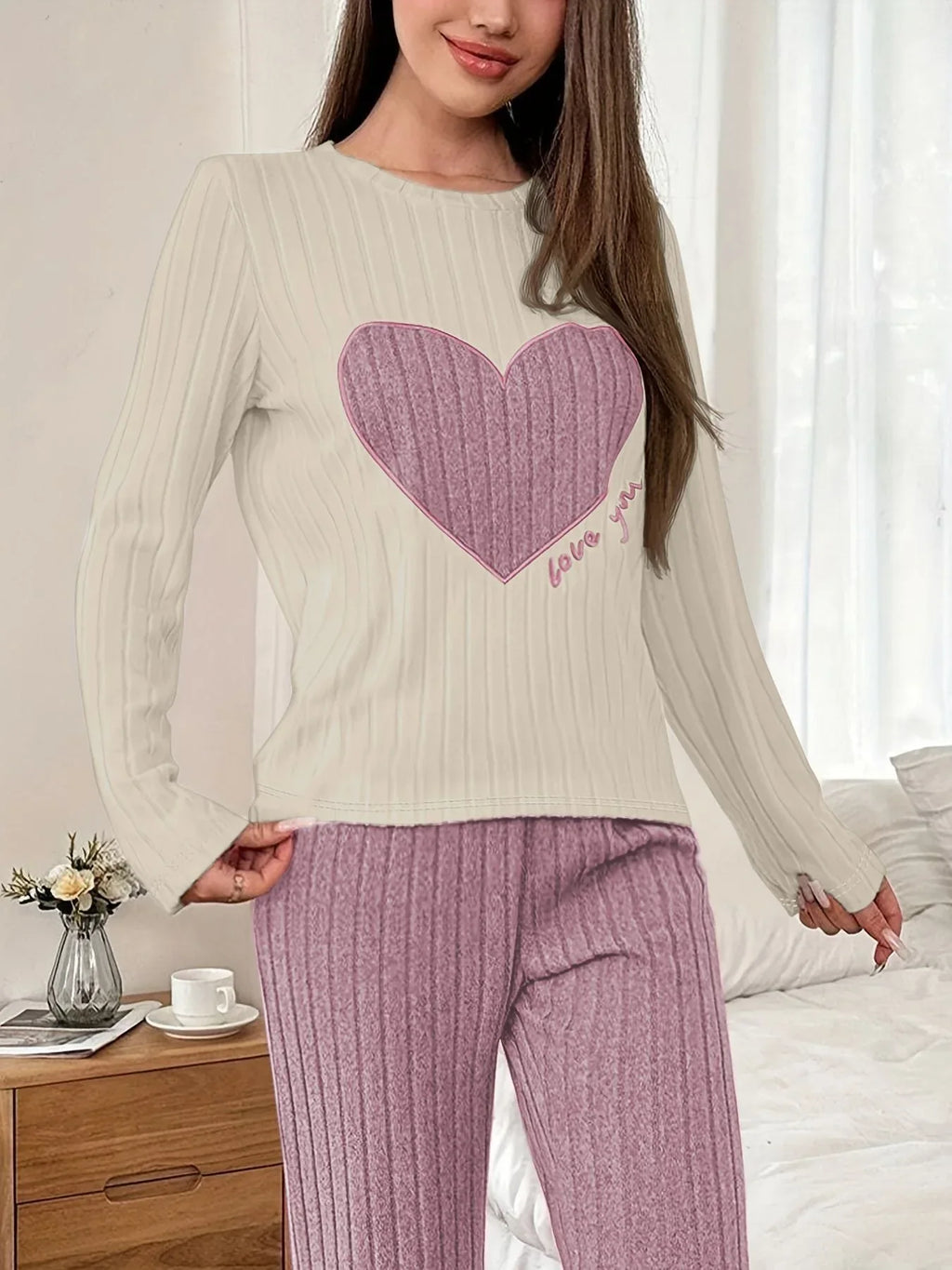 Woman wearing a cream long-sleeve ribbed pajama top with a pink heart graphic and lilac ribbed pajama pants, standing in a bedroom next to a nightstand.
