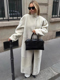 Woman in a long cream sherpa coat and white pants, holding a black crocodile-embossed handbag.
