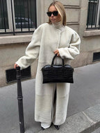 Woman in a long cream sherpa coat and white pants, holding a black crocodile-embossed handbag.
