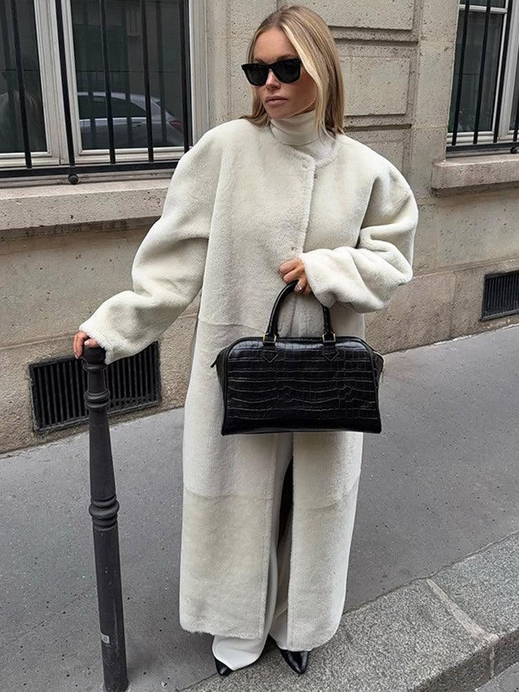 Woman in a long cream sherpa coat and white pants, holding a black crocodile-embossed handbag.