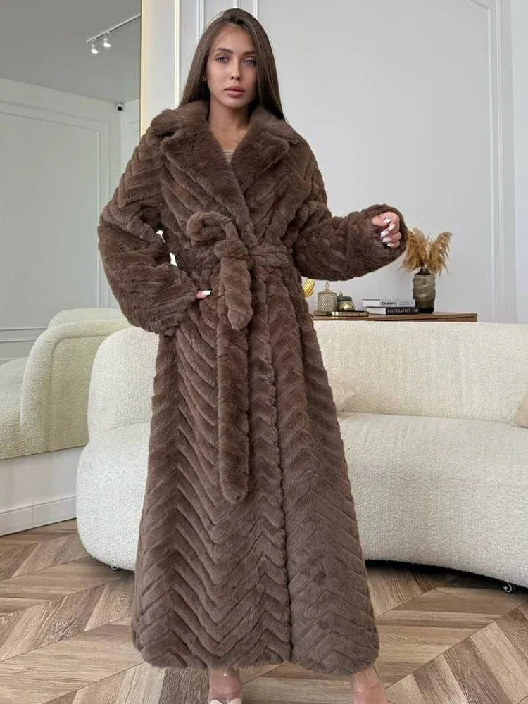 Woman wearing a long, brown faux fur coat with a chevron pattern and a belted waist, posing indoors.
