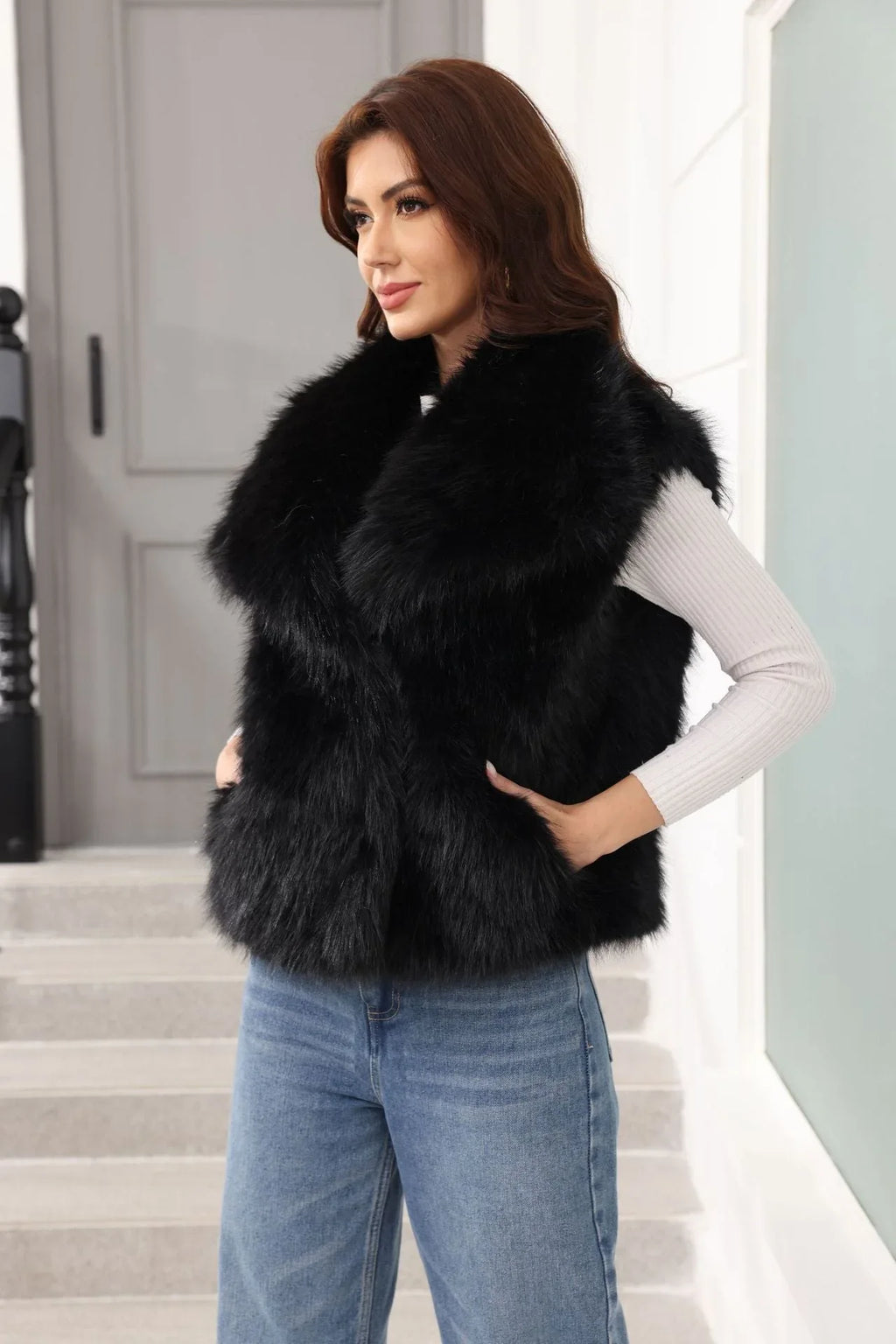 Woman wearing a black faux fur vest and blue jeans, with hands on hips, looking to the side.