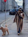 Full shot of a woman in a long faux fur coat walking her dog on a cobblestone street.