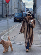 Full shot of a woman in a long faux fur coat walking her dog on a cobblestone street.