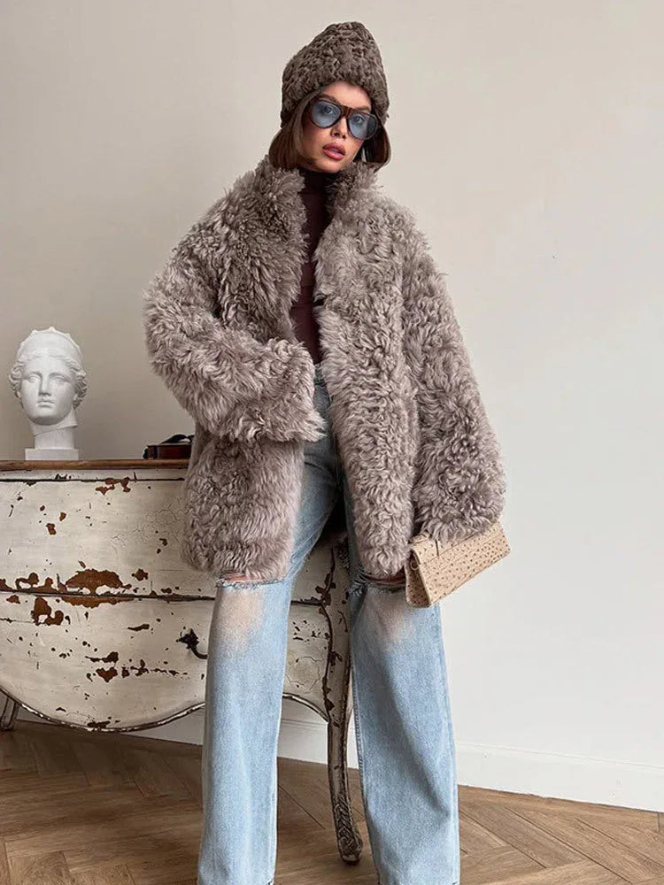 Woman wearing a grey curly faux fur oversized coat with a brown turtleneck and light blue ripped jeans, holding a cream clutch.