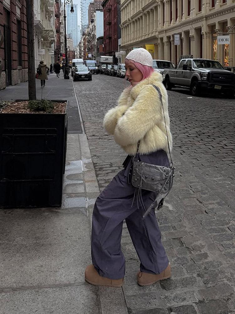 Woman wearing a cream faux fur cropped jacket with a lapel collar and long sleeves, paired with grey pants and a white knit beanie, walking on a cobblestone street.