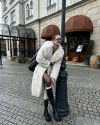 Woman leaning against a lamppost, wearing a white faux fur coat and black boots.