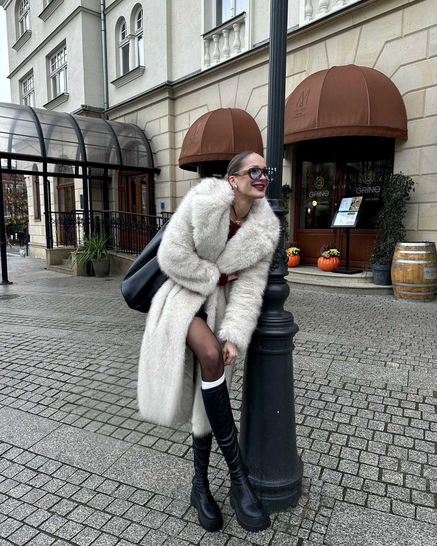 Woman leaning against a lamppost, wearing a white faux fur coat and black boots.