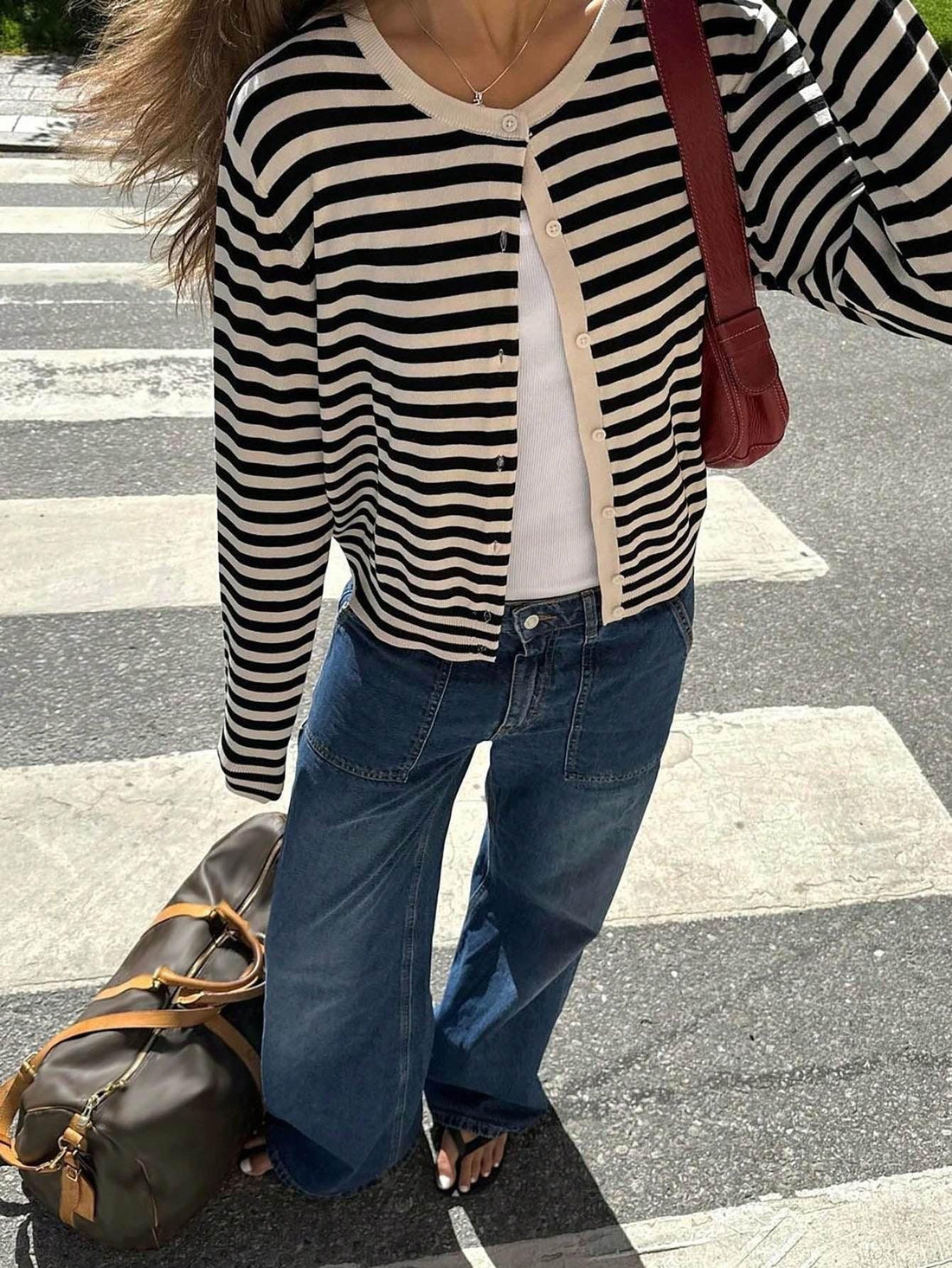 Woman wearing a black and white striped cardigan with a white tank top and blue wide-leg jeans standing on a crosswalk.