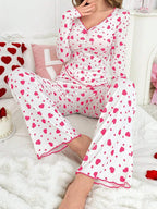 Woman sitting on a bed wearing a white long-sleeved pajama top and pants with a pink heart print, with her legs crossed.