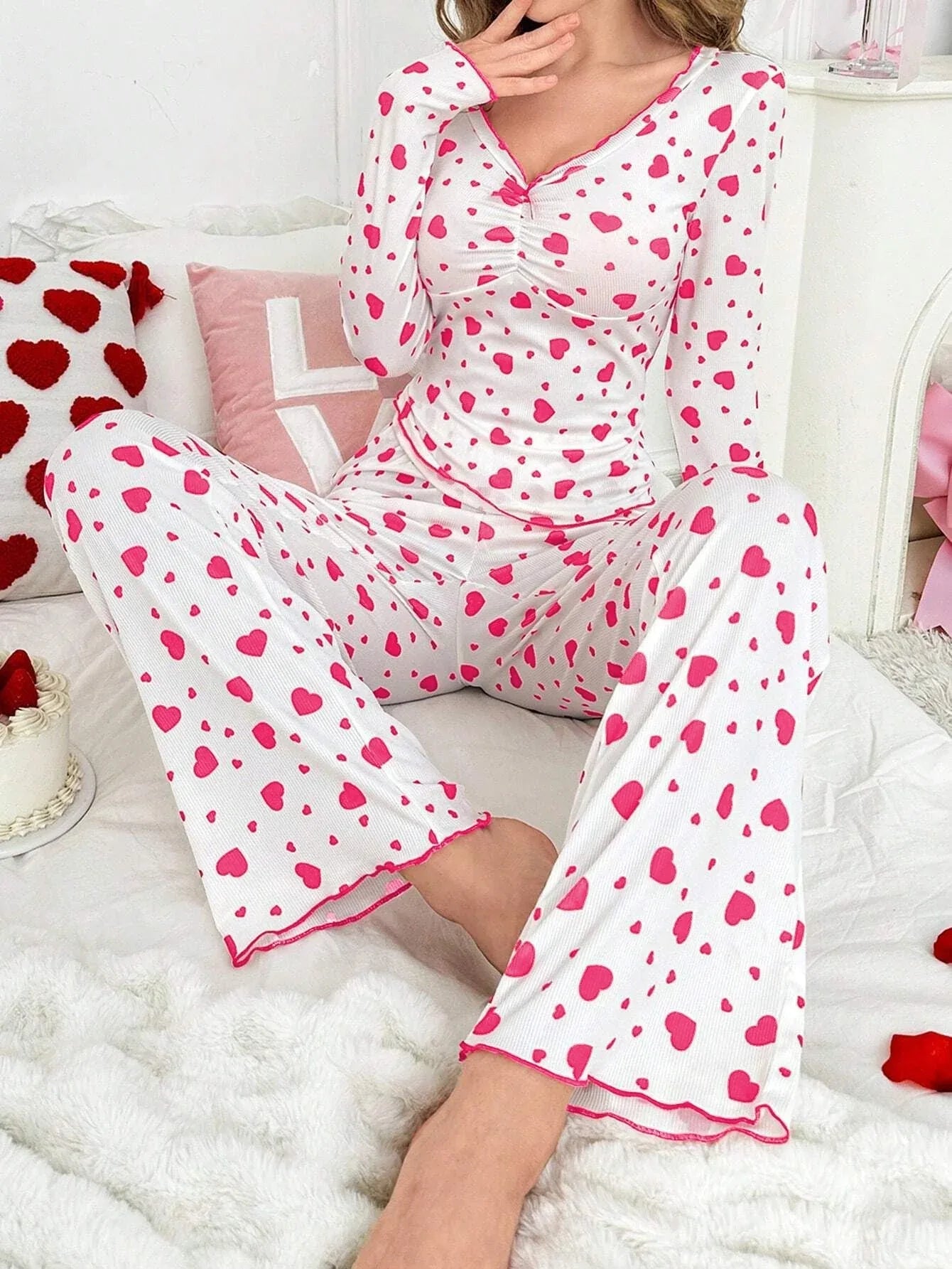 Woman sitting on a bed wearing a white long-sleeved pajama top and pants with a pink heart print, with her legs crossed.
