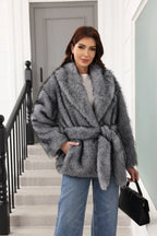 Woman in a grey faux fur coat with a belt, posing with a handbag.