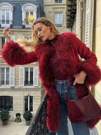 Woman in a red suede coat with faux fur trim and cuffs, standing on a balcony with a red handbag.