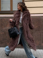 Woman wearing a long brown shaggy faux fur coat and blue jeans, holding a black handbag.