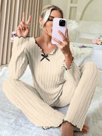 Woman taking a selfie on a bed while wearing a cream-colored ribbed knit loungewear set with black trim and a black bow.