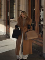 Woman in a long camel coat with shearling trim and white pants, holding shopping bags and a brown handbag.