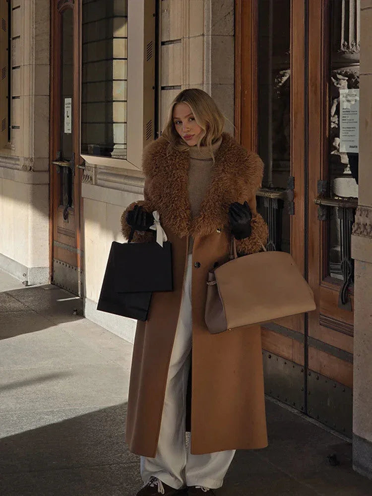 Woman in a long camel coat with shearling trim and white pants, holding shopping bags and a brown handbag.