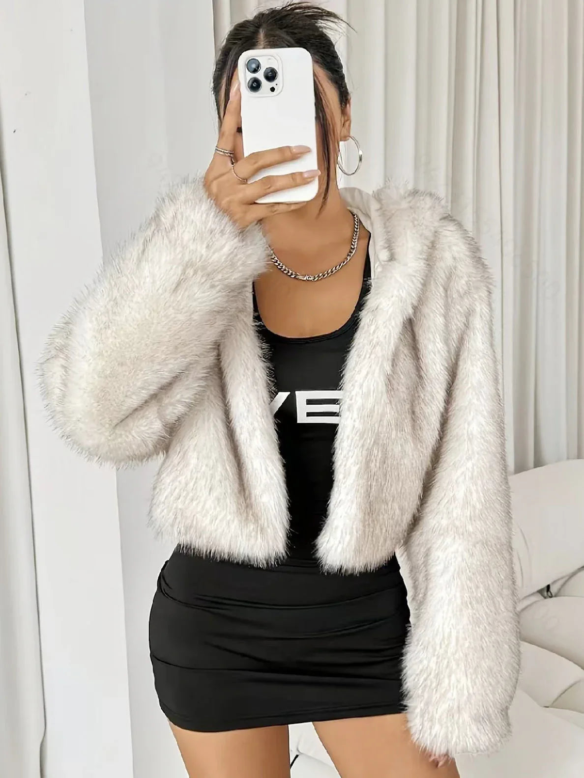 Woman wearing a white faux fur cropped hooded jacket over a black dress, holding a phone Main image