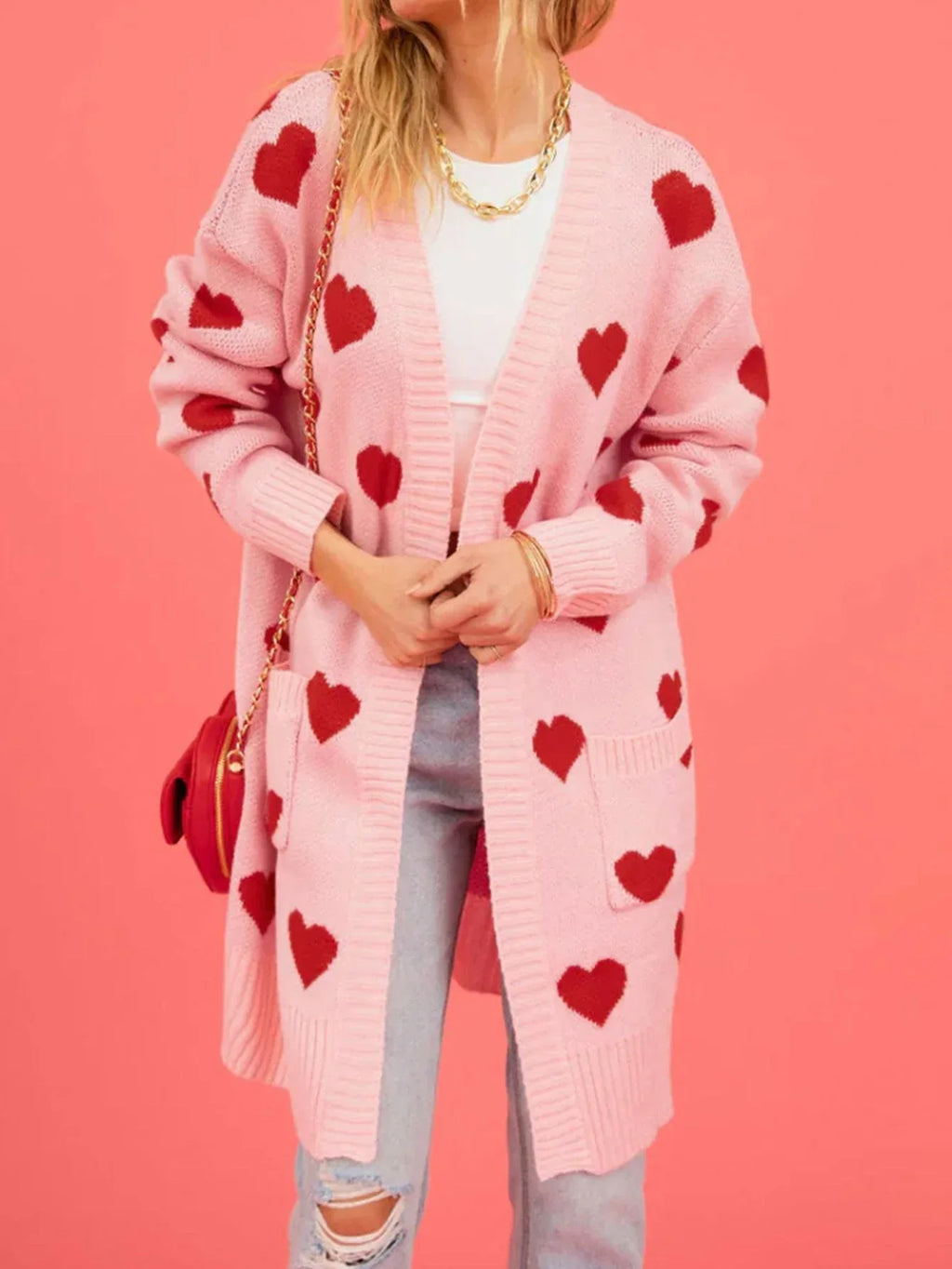 Woman wearing a pink longline knit cardigan with a red heart pattern and two front pockets.