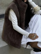 Woman sitting down wearing a brown faux fur vest, white long-sleeved shirt, and white pants.