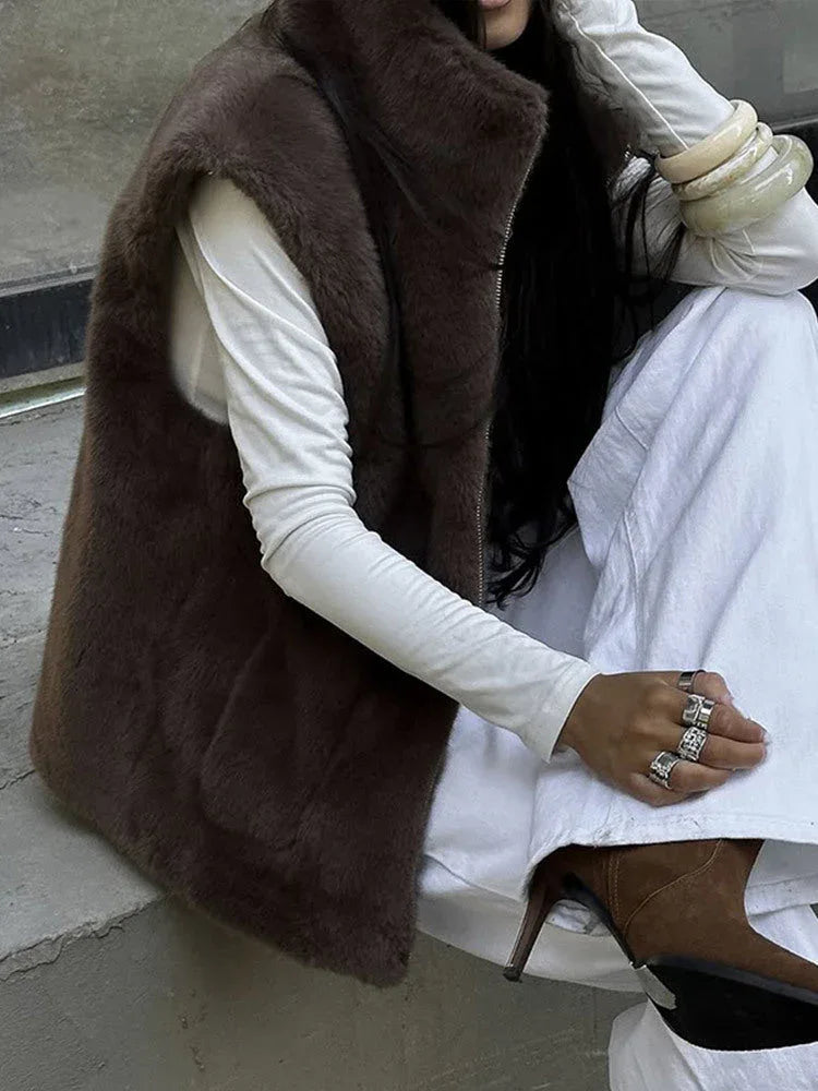 Woman sitting down wearing a brown faux fur vest, white long-sleeved shirt, and white pants.