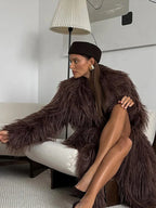 Woman sitting on a white sofa wearing a long brown shaggy faux fur coat and a brown beret.
