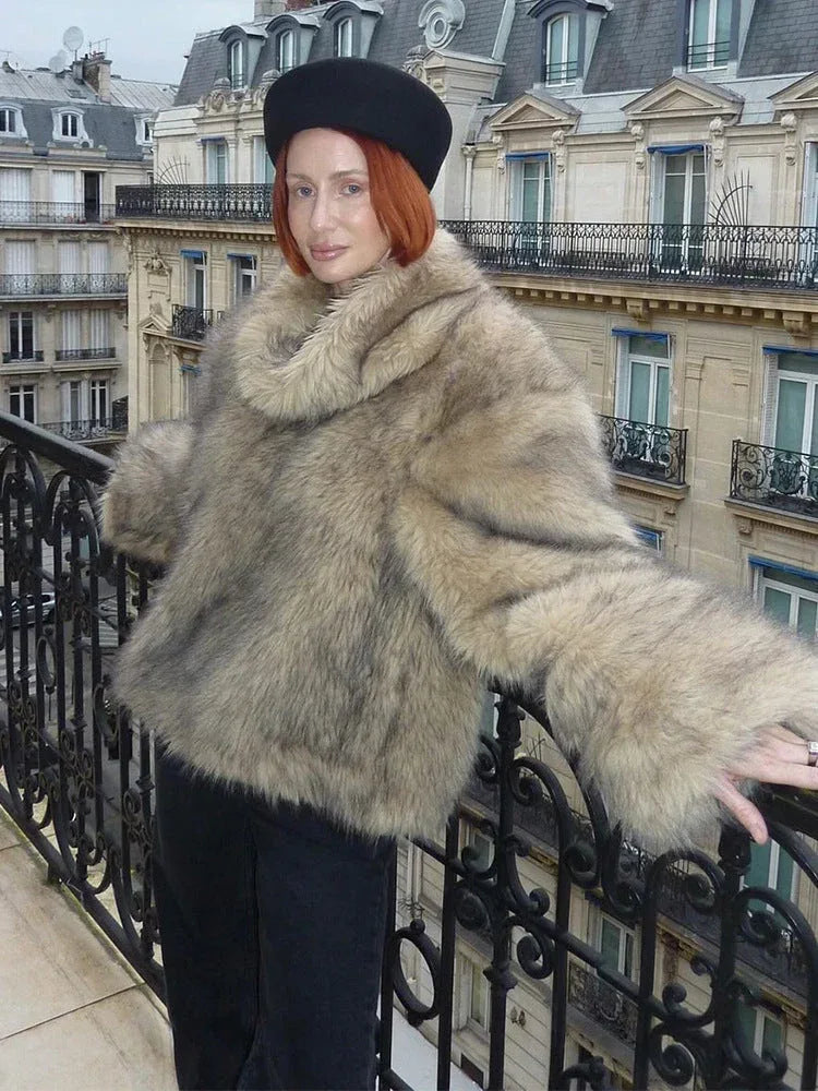 Woman wearing a light brown faux fur jacket with a wide collar and full-length sleeves, standing on a balcony.