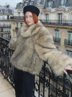 Woman wearing a light brown faux fur jacket with a wide collar and full-length sleeves, standing on a balcony.