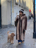 Woman in a brown and beige striped faux fur coat holding a coffee cup and walking a dog.
