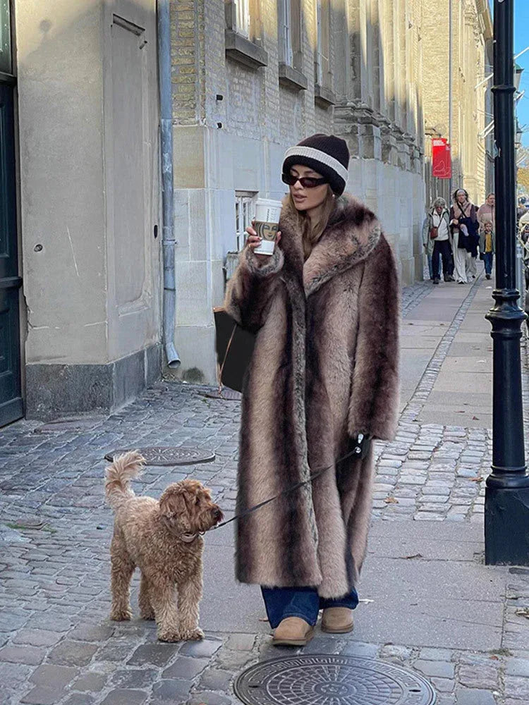 Woman in a brown and beige striped faux fur coat holding a coffee cup and walking a dog.