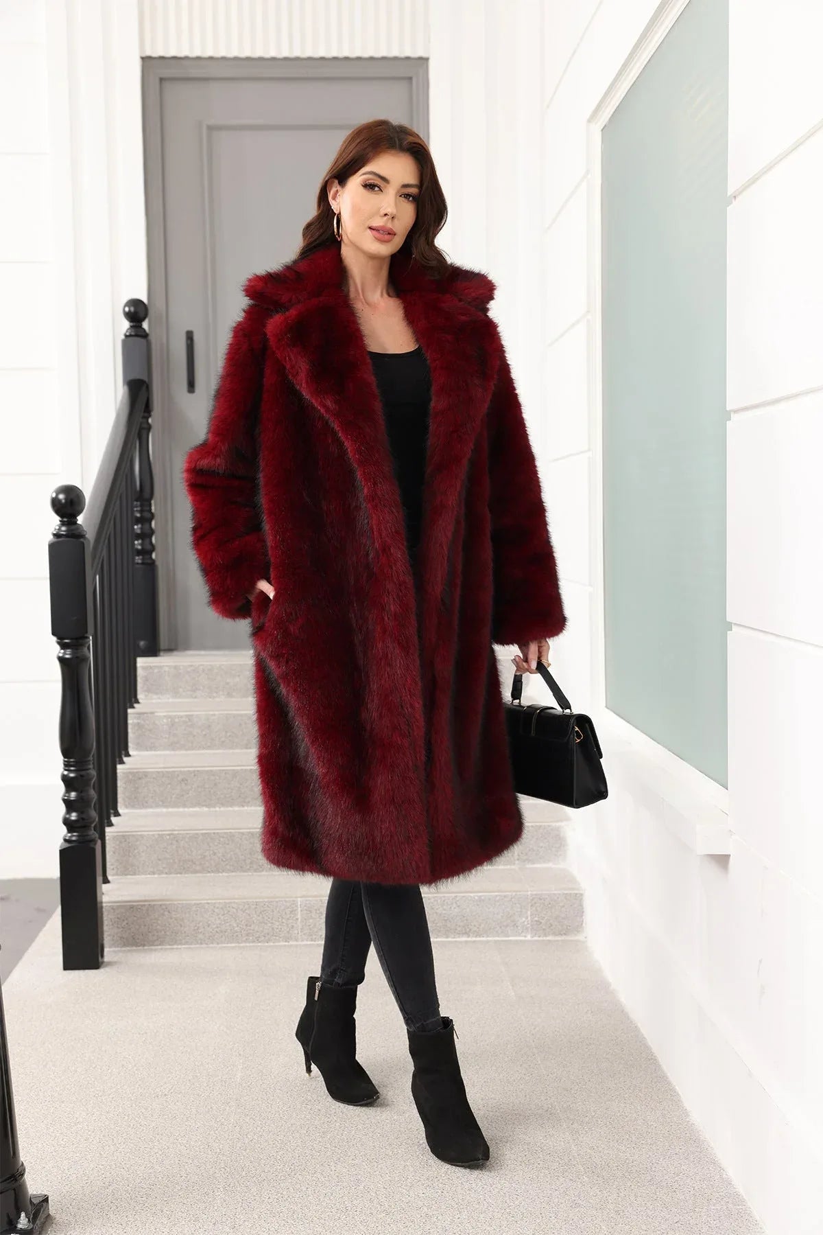 Woman in a dark red faux fur coat and black pants, holding a black handbag, standing on stairs.
