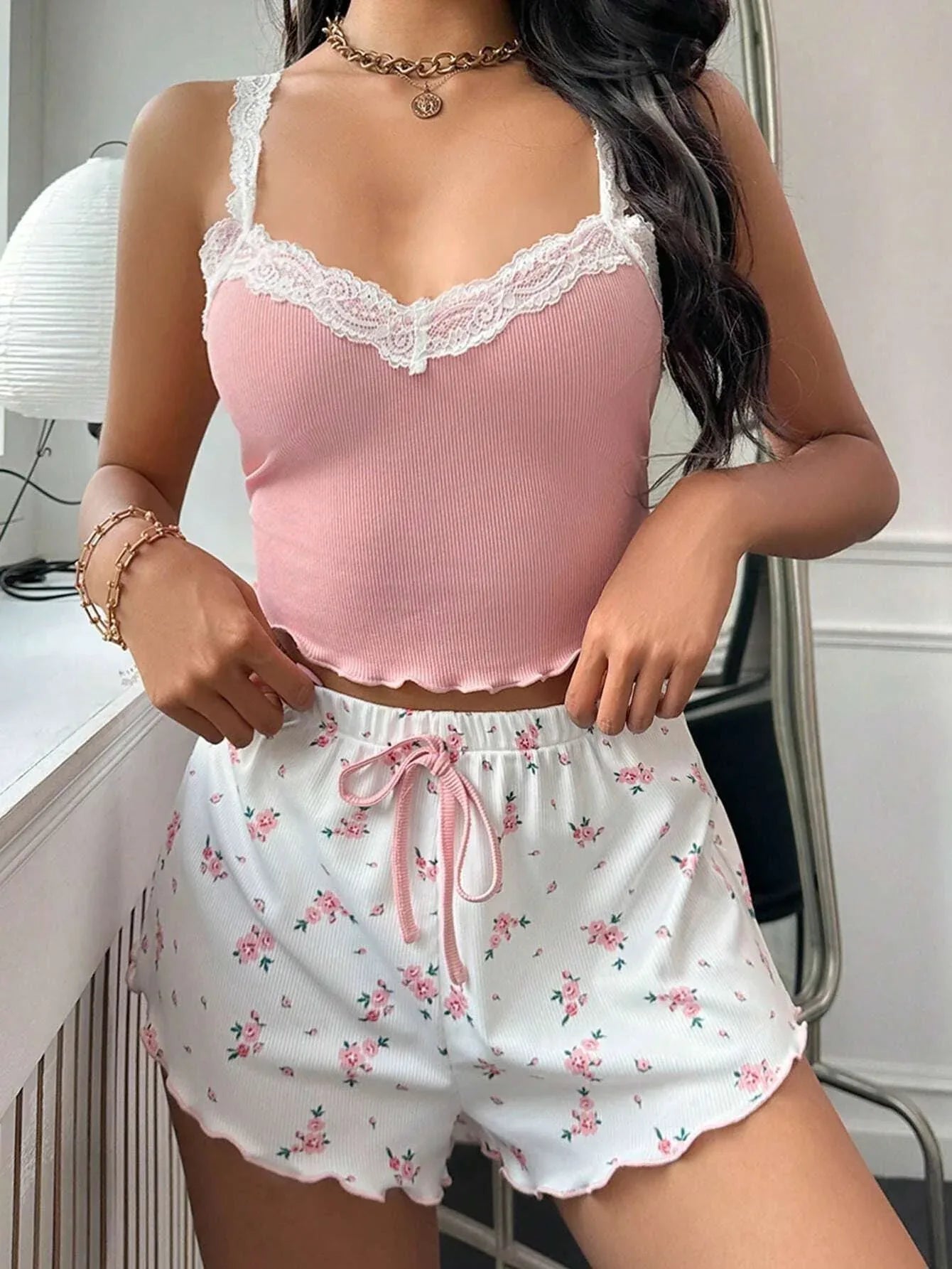 Woman adjusting the waistband of white floral print shorts with pink trim and a pink drawstring while wearing a pink ribbed knit camisole with white lace trim.