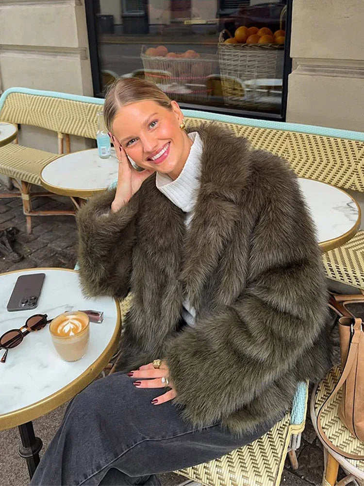 Woman wearing a green faux fur jacket and white turtleneck sitting at an outdoor cafe table. Main image