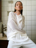 Woman wearing a white sheer knit sweater and white pants.