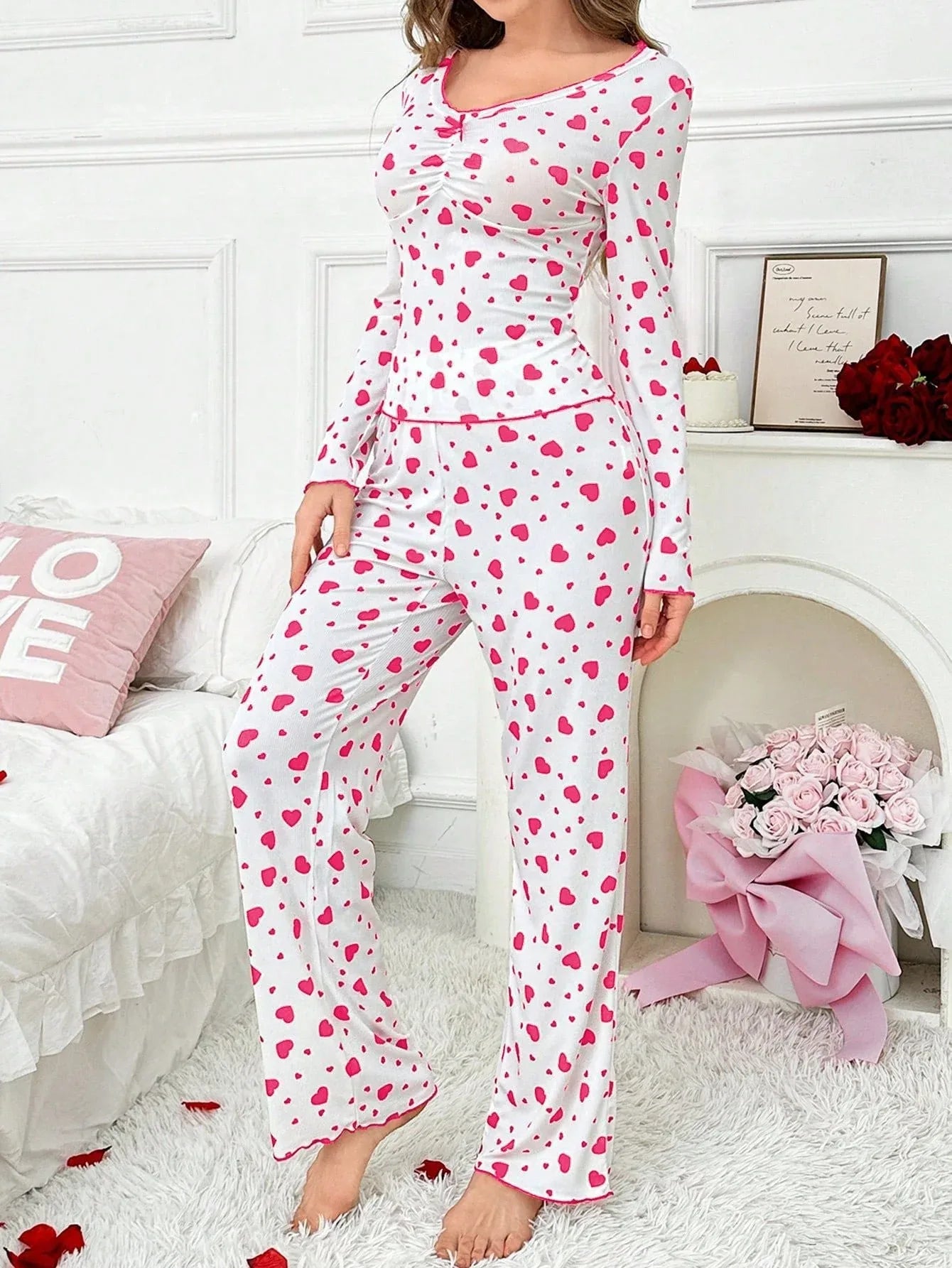 Woman wearing a white long-sleeved pajama top and pants with a pink heart print, standing in a bedroom. Main image