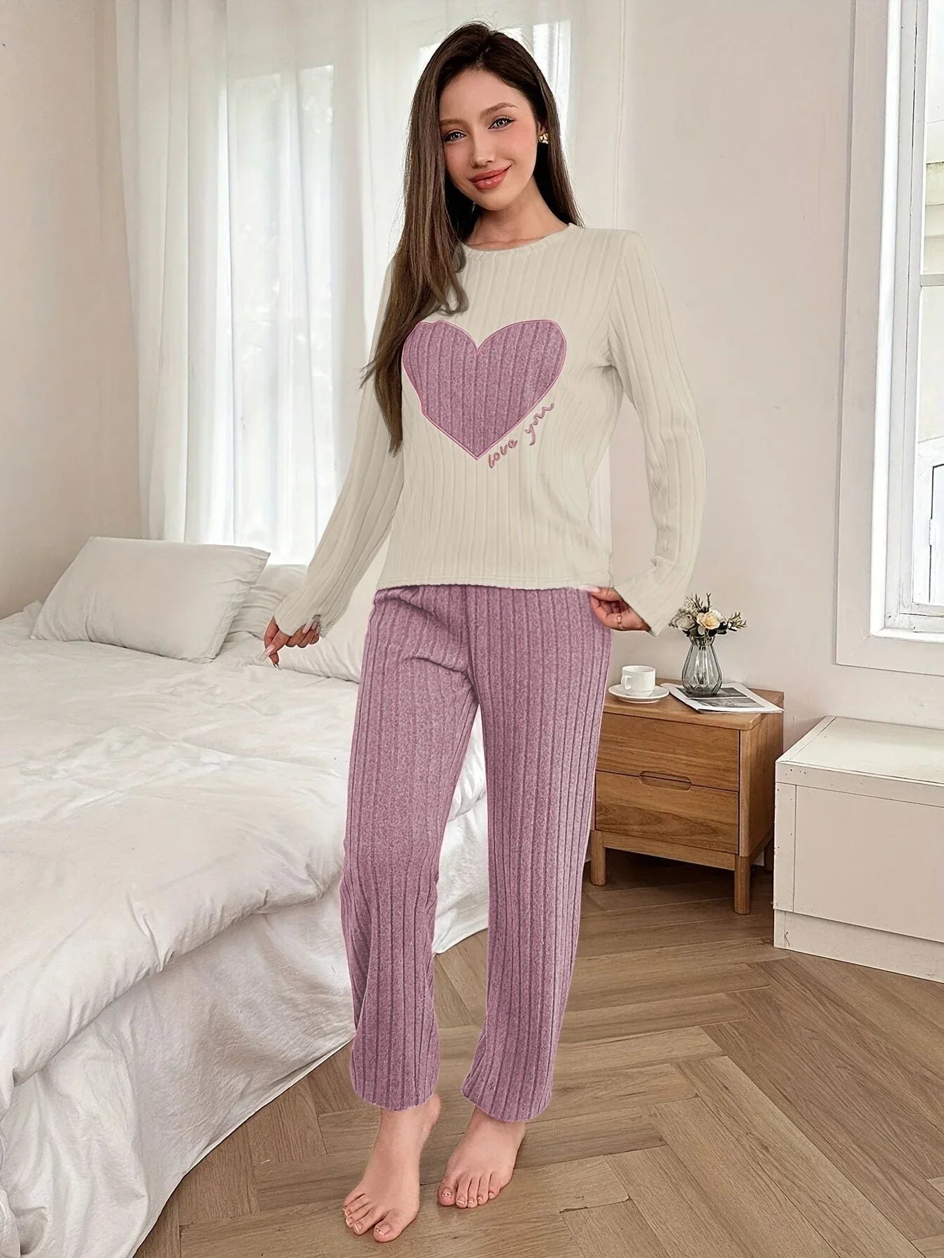 Woman wearing a cream long-sleeve pajama top with a large purple heart graphic and the words 'love you' printed on it, paired with purple ribbed full-length pajama pants, standing in a bedroom.
