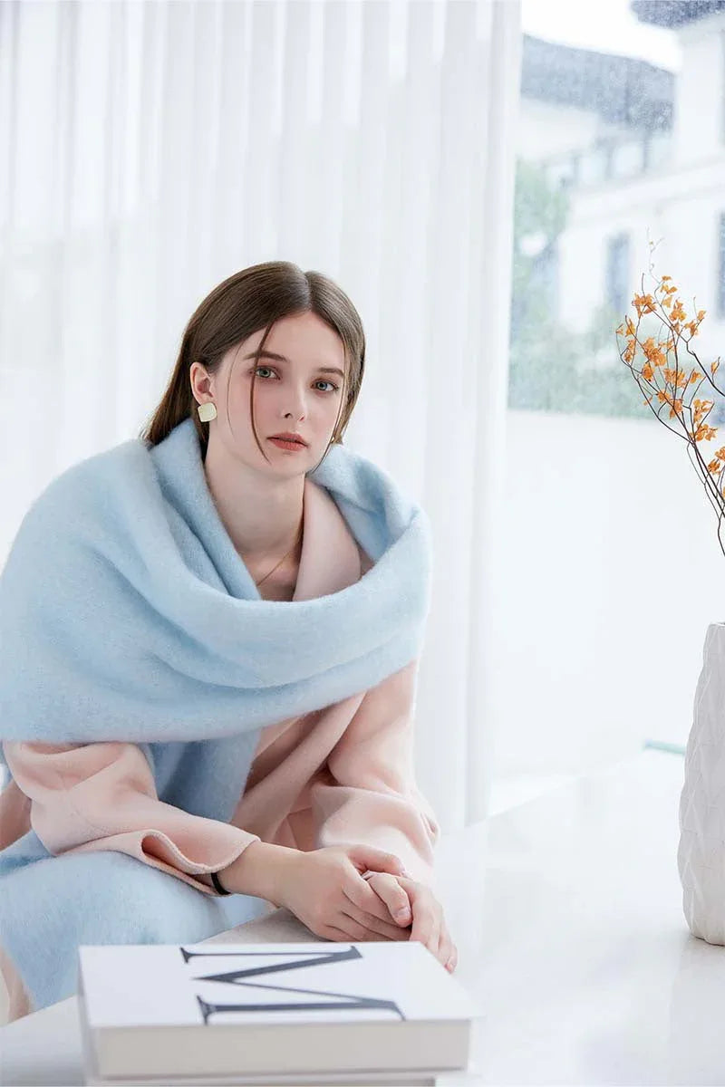 Woman wearing a light blue oversized scarf draped over a pink coat, seated indoors.