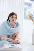 Woman wearing a light blue oversized scarf draped over a pink coat, seated indoors.
