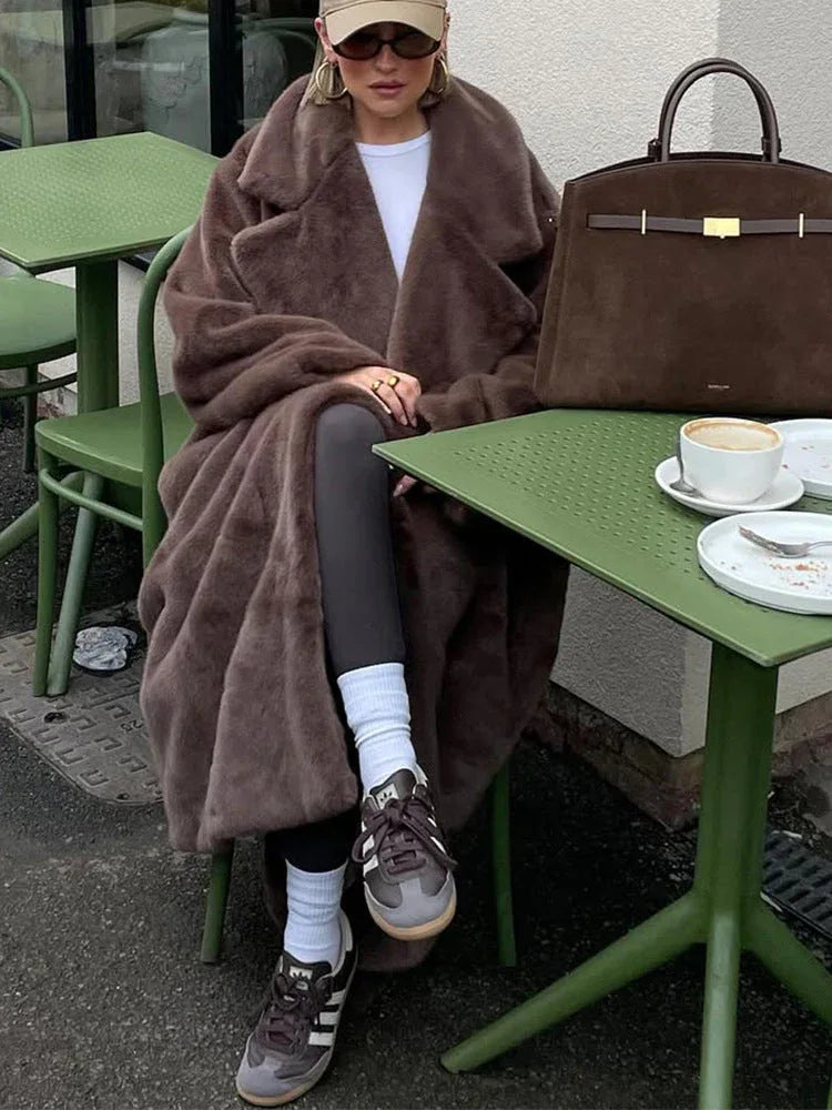 Woman with a brown faux fur coat, sunglasses, and a baseball cap sitting at a table.
