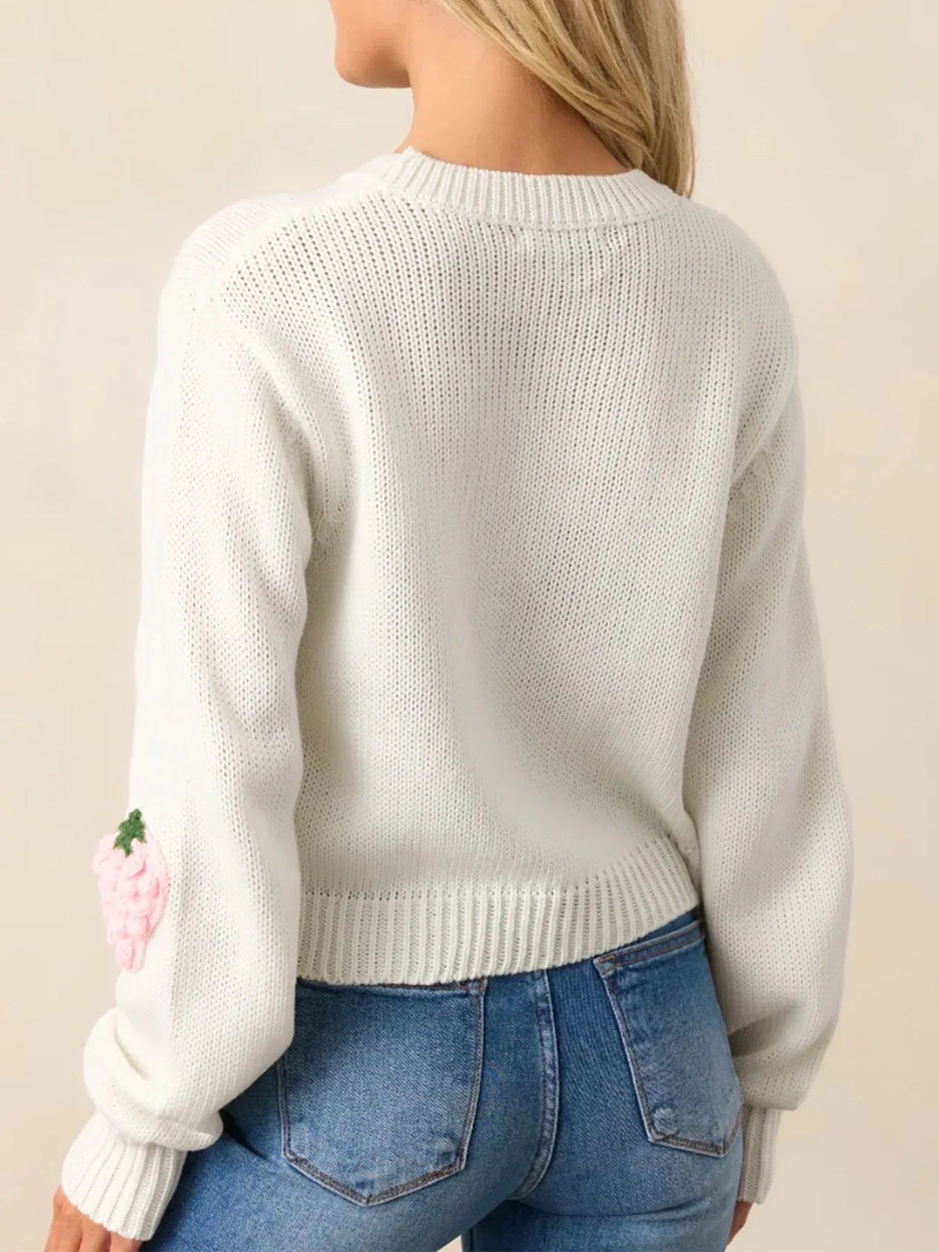 Rear view of a woman wearing a white cropped knit sweater with a strawberry applique on the sleeve, paired with blue denim jeans.