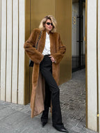 Woman wearing a long brown faux fur coat over a white shirt and black pants.