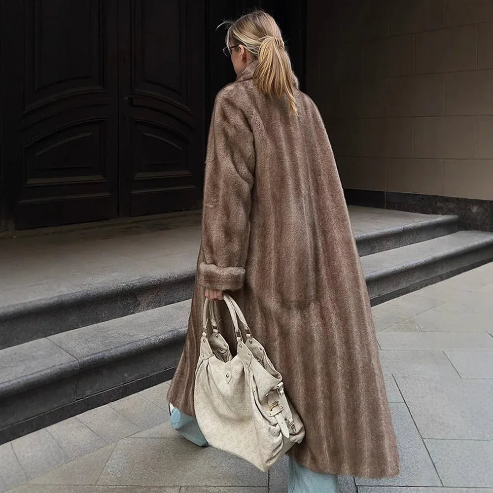 Rear view of a woman wearing a long brown faux fur coat, white t-shirt, and light blue wide-leg jeans, holding a cream-colored handbag.