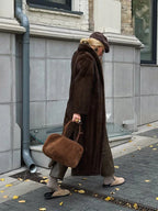 Woman walking in a long brown faux fur coat, taupe knit sweater, taupe trousers, taupe beret, sunglasses, and tan slip-on shoes, holding a brown suede handbag.