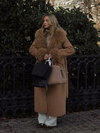 Woman in a long camel coat with shearling trim and white pants, holding shopping bags.