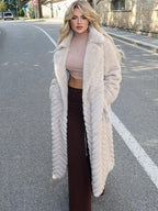 Woman wearing a long, cream-colored faux fur coat with a chevron pattern and a belted waist, standing outdoors on a street.