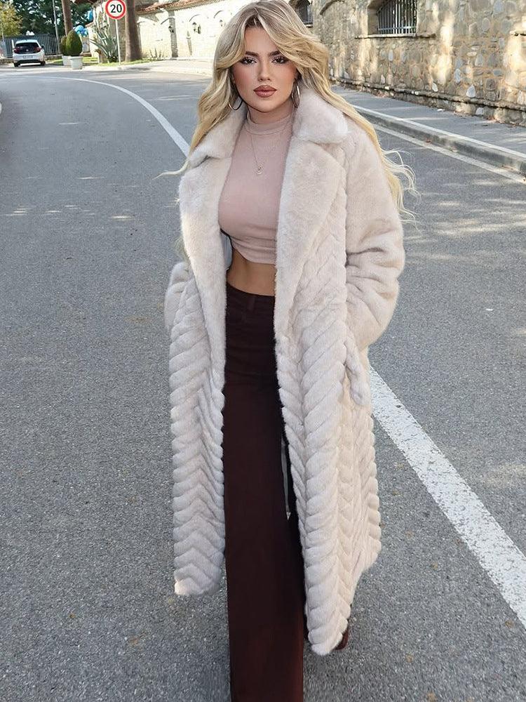 Woman wearing a long, cream-colored faux fur coat with a chevron pattern and a belted waist, standing outdoors on a street.