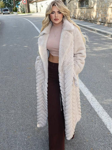 Woman wearing a long, cream-colored faux fur coat with a chevron pattern and a belted waist, standing outdoors on a street.