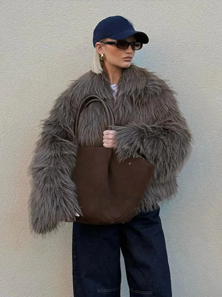 Woman wearing a brown shaggy faux fur jacket, dark wash wide-leg jeans, a navy baseball cap, and sunglasses. She is holding a large brown tote bag.