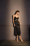 Woman wearing a black satin slip dress with lace bust detail and a matching black robe, standing against a grey wall.